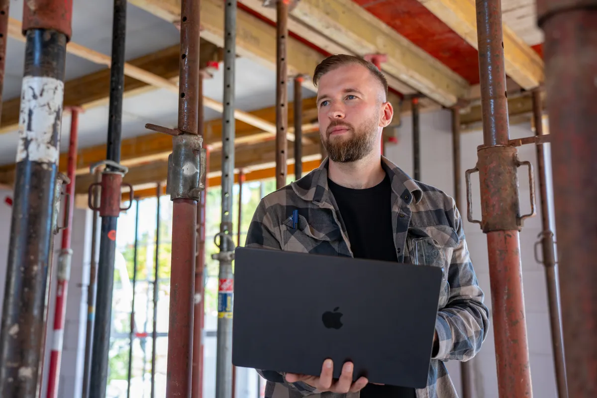 Marvin Jennrich steht mit aufgeklapptem Laptop zwischen Deckenstützen in einem Rohbau